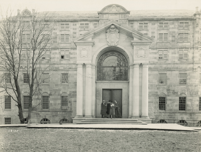 The front entrance of the Memorial Union into Gold Star Hall and focuses on the entryway. There are four people standing on the front steps, in front of the large bronze doors. A boardwalk runs along the front of the building, and makes a wider platform around the entrance and stone steps. Another woman is walking on the boardwalk toward the entrance, and being in motion, appears as a blurred, ghostly image.