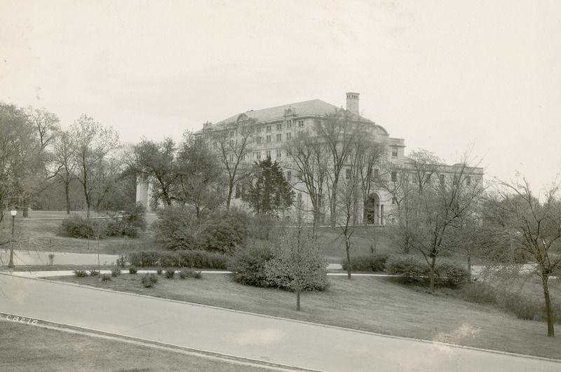 This distance image shows the Memorial Union through trees and shrubbery in the foreground. The side (west) and Gold Star Hall entrances are visible through the trees, and the roofline and one chimney are prominent against the sky. A paved road and sidewalk are in the foreground.