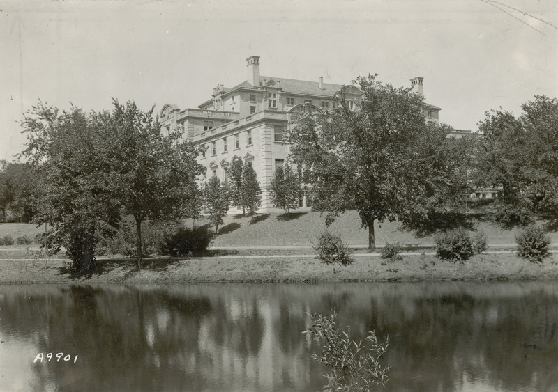 This image focuses on one corner, the southwest, of the Memorial Union with Lake LaVerne down an embankment and in the foreground. Trees and shrubbery partially hide the building, but the photograph is composed to emphasize the roof levels, chimneys, balconies and pediments of the building.