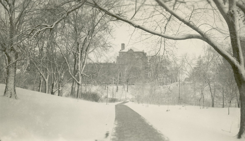 A sidewalk leads through a snow covered lawn to the north side of the Memorial Union. The Union appearing in the background is partially concealed the trees and bushes.