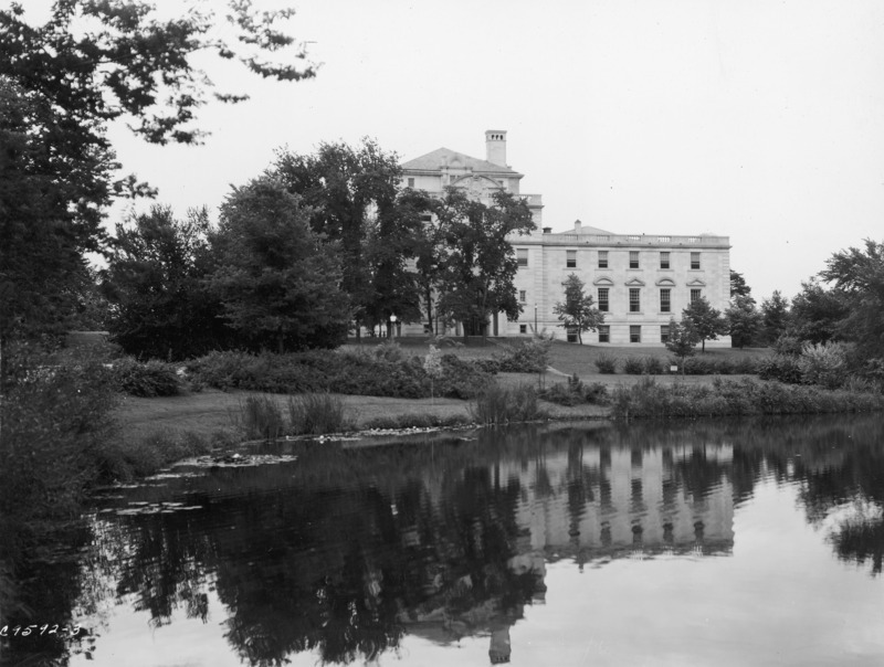 The east facade of the Memorial Union is partially hidden from view by trees. Both the Union and trees are reflected in Lake LaVerne.