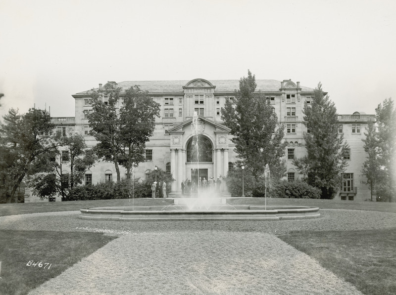 This 1937 photograph shows the front (north) of the Memorial Union, viewed from across the fountain. The fountain has several water jets going upward. A group of several people is standing on the steps in front of the entrance to Gold Star Hall. Taken from a distance, this photograph illustrates many of the Union's details such as the columns and carvings around the entrance to Gold Star Hall, the balustrades along the roof edge, the faux balconies under some windows, the pediments and the slate roof.