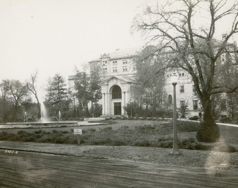 This 1938 image taken from north and only slightly west of the Gold Star Hall (front) entrance to the Memorial Union has foreground an unpaved road that crosses in front of the fountain and defines the Union's property. A lamppost and a No Parking sign are along the edge of the road. The Gold Star Hall entrance is centered and visible are its architectural details including columns, carvings, and the arch with arched window.