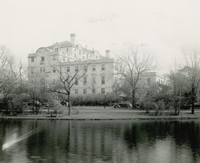 This 1941 photograph is of the west side of the Memorial Union, seen from across Lake LaVerne. Several cars are parked and people are walking along a road between the lake and the Union. Plantings partially obscure the lower story of the Union, but the upper floors and distinctive roof lines, roof levels, balustrades, chimneys and pediments are visible.