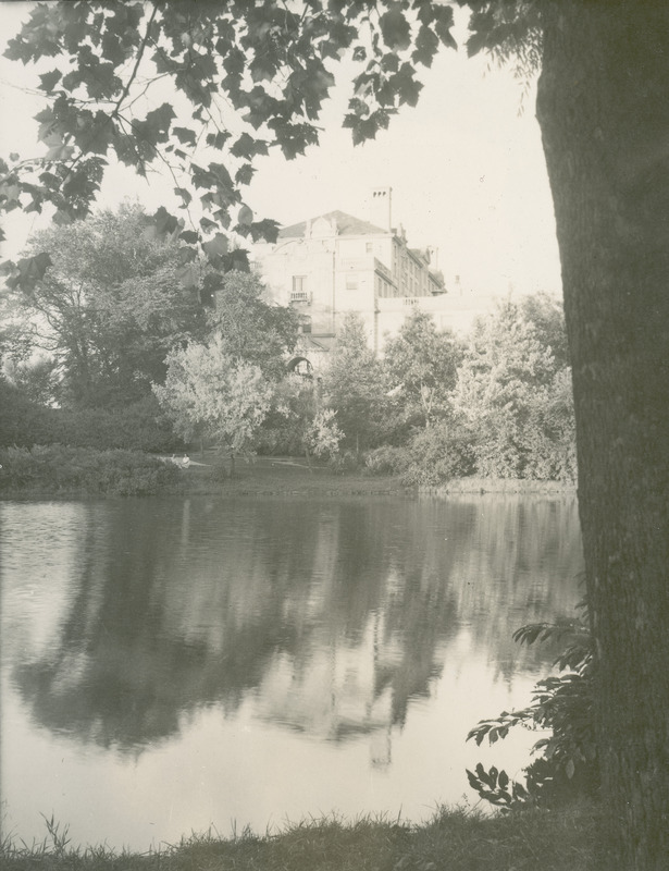 This 1944 photograph of the Memorial Union's west side shows mainly the roof lines, chimneys, pediments, a balcony around a window above the entrance, and the windows on the upper floors. Lake LaVerne is in the foreground, and plantings and trees hide most of the Union building. Two women are sitting on the far bank of the lake.