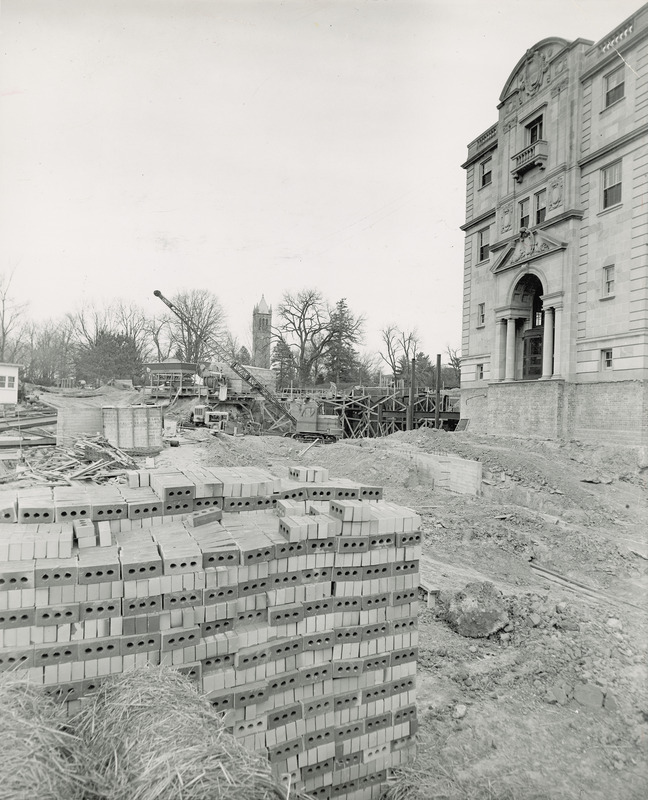 This 1950 photograph of the Memorial Union shows the excavated construction area for what will become the west terrace. A new foundation wall is near the center. A large stack of bricks is in the foreground and a crane, steel framework for the northwest addition, forms and cement mixing machinery are in the distance. In the far distance is the Campanile.