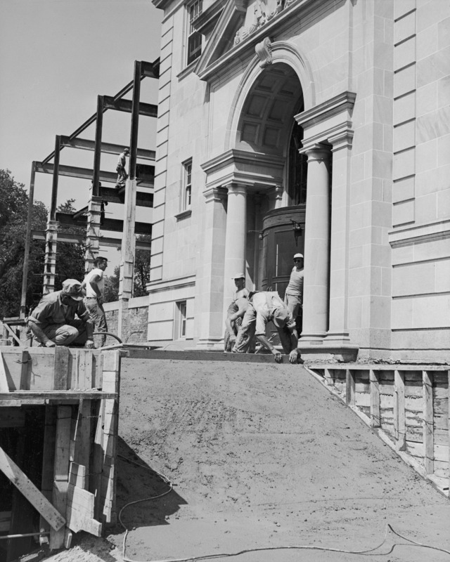 This 1951 photograph shows a concrete pour in progress on the new west terrace, outside the west entrance to the Memorial Union. The steel framework for the northwest addition is seen beyond the corner of the building. Five workmen are involved the pour and one is walking on a girder.