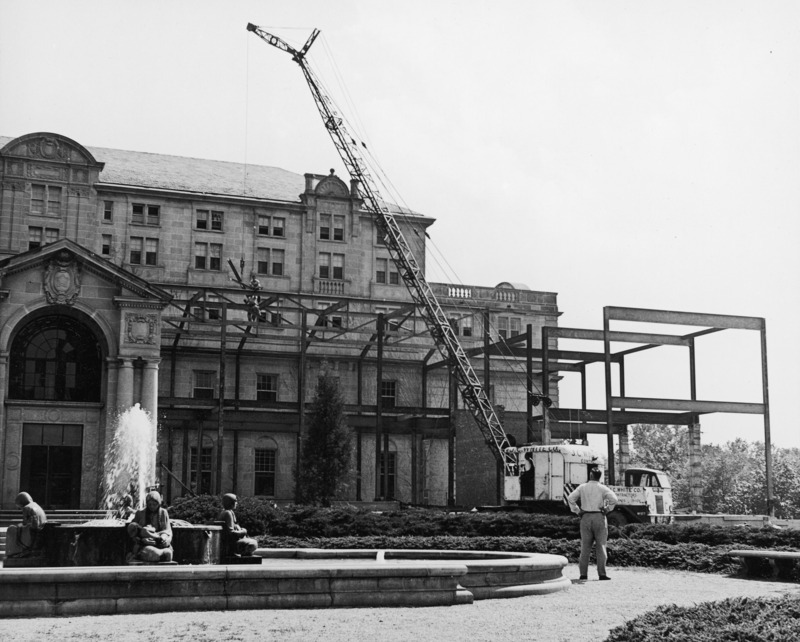 This 1951 photograph is of the steel framework of the northwest addition to the Memorial Union. A crane is lifting a beam while two workmen guide it into place. The Four Seasons Fountain is in the foreground with the Gold Star Hall entrance to the left. A lone spectator watches the work.