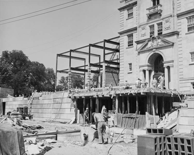 This photograph shows work progressing on the west terrace of the Memorial Union in 1951. Supports hold up the upper deck outside the building's west entrance. Several workmen are doing various tasks around the site, one in the lower center is mixing mortar. The terrace wall is partially complete and the framework of the northwest addition is in the center of the image.