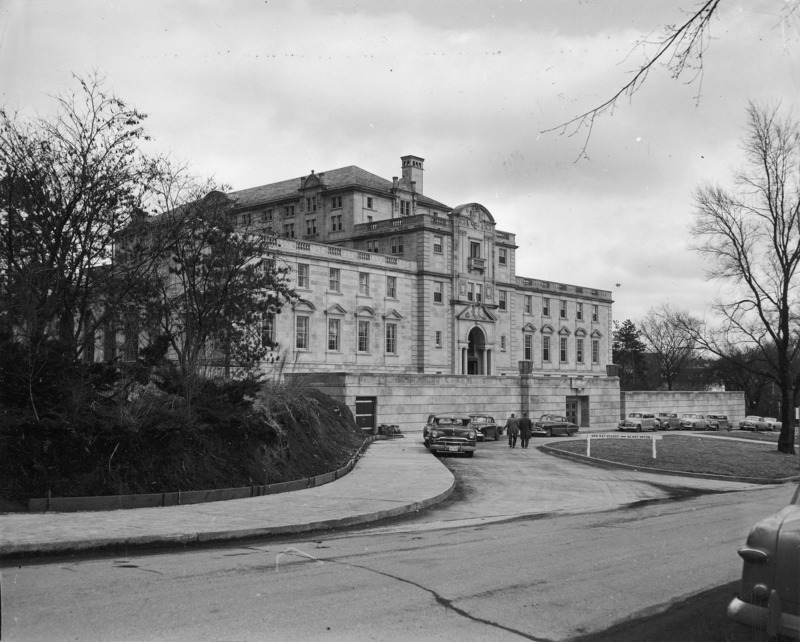 This 1953 photograph shows the entire west side and terrace of the Memorial Union from street level. In it are easily seen many of the Union's unique architectural details including the pediments over the windows and in the roofline, balustrades, chimneys and balconies. Several cars are parked along the curved street, and two people are walking towards the street level entrance. The hill to the left is bare and un-landscaped, and a temporary barrier runs along its foot to keep dirt from spilling onto the sidewalk.