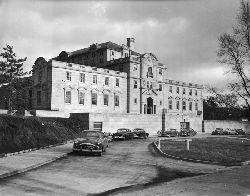 This 1953 photograph shows the entire west side and terrace of the Memorial Union from street level. In it are easily seen many of the Union's unique architectural details including the pediments over the windows and in the roofline, balustrades, chimneys and balconies. Several cars and a bicycle are parked along the curved street. The hill to the left is bare and un-landscaped, and a temporary barrier runs along its foot to keep dirt from spilling onto the sidewalk.