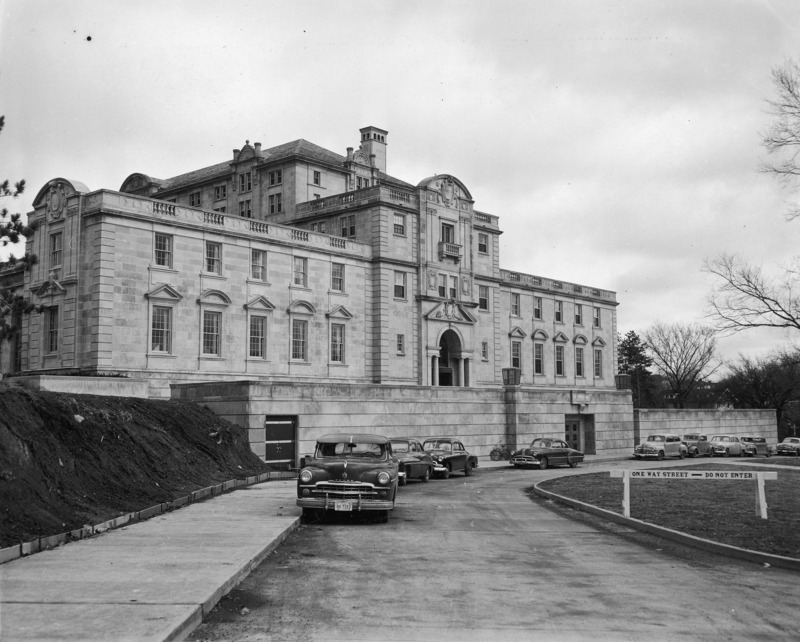 This 1953 photograph shows the entire west side and terrace of the Memorial Union from street level. In it are easily seen many of the Union's unique architectural details including the pediments over the windows and in the roofline, balustrades, chimneys and balconies. Several cars and a bicycle are parked along the curved street. The hill to the left is bare and un-landscaped, and a temporary barrier runs along its foot to keep dirt from spilling onto the sidewalk.