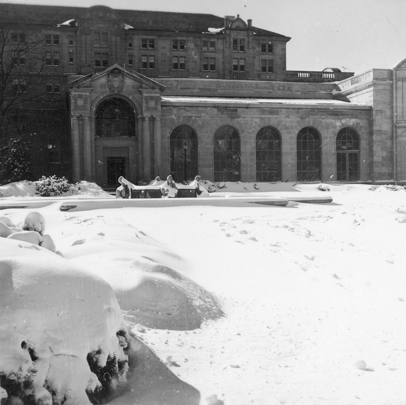 This 1953 winter photograph focuses on the north facade of the new northwest addition, west of the Gold Star Hall entrance, to the Memorial Union. Snow covers the ground, shrubbery and Four Seasons Fountain in the foreground. The five large arched windows in the addition repeat the shape of the window in the entrance.