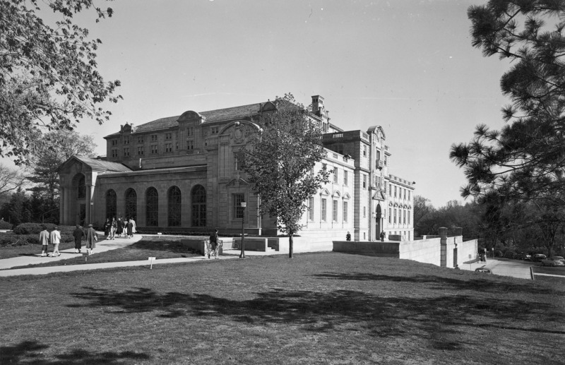 This 1954 photograph of the front (north) and (west) side of the Memorial Union has several people walking on the curved sidewalk towards Gold Star Hall and on the west terrace. The west side of the building is in bright sunlight and the front (north) slightly in shadow. The photograph highlights the northwest addition, with the whole building set against a clear sky. Tree branches and an expanse of lawn are in the foreground.