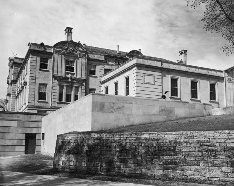 This 1954 photograph of the southwest (back) corner of Memorial Union is taken from street level and shows part of the south wing. Prominent are a stone retaining wall, the walls of the lower terrace and the west corner of the south wing. The back (south) and the side (west) walls of the original building rise on the left. Corners are emphasized, as is the layering effect in which the building and landscaping rise above the street.