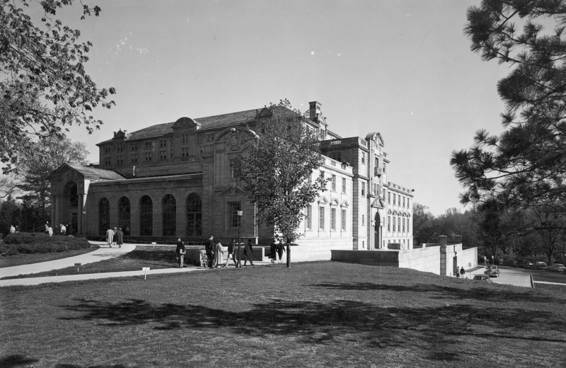 This 1954 photograph of the front (north) and (west) side of the Memorial Union has several people walking on the curved sidewalk towards Gold Star Hall, and towards the west terrace. The west side of the building is in bright sunlight and the front (north) slightly in shadow. The photograph highlights the northwest addition, with whole building set against a clear sky. Tree branches and an expanse of lawn are in the foreground.