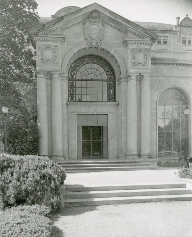 This 1955 photograph is of the outside entrance to Gold Star Hall in the front (north) of the Memorial Union. Decorative details of the entrance include the four attached columns with Doric-style capitals, decorative carvings incorporating grotesques, rosettes, and the five-pointed star in the peak of the pediment, and the large arched window. A revolving door is in the doorway which has been modified from its original size.