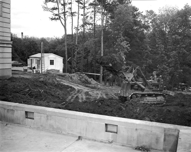 This 1957 photograph taken from the terrace is of the beginning excavations for what will be the bookstore and Sun Room in the Memorial Union. A workman in a skid shovel with the shovel full of dirt and raised high waits and looks towards a dump truck that is emptying its load just beyond some trees. The earth around the skid shovel is stripped bare and gouged, tangled with tree roots and shows tire tracks under the raised shovel. The wall of the terrace is in the foreground. A white construction cabin with a table and various wood and sawhorses around it is beyond the back (south) corner of the Union.