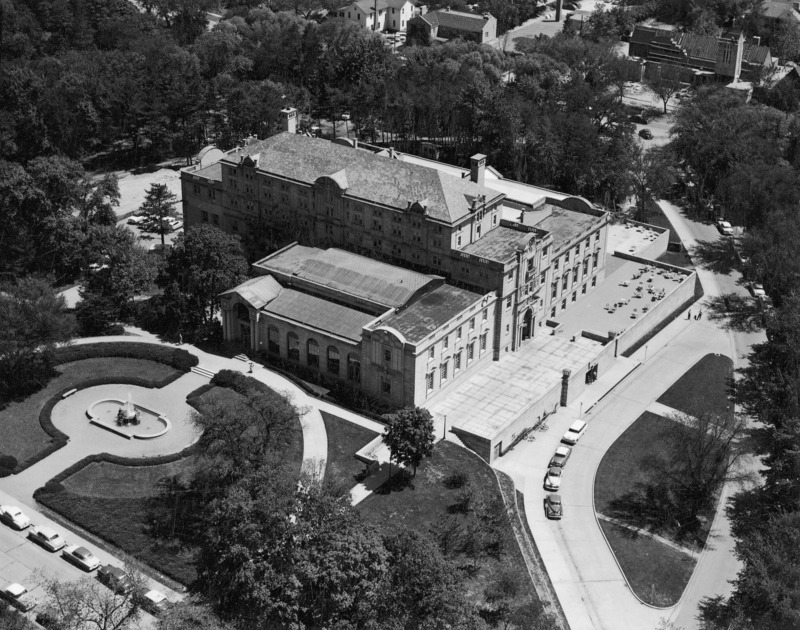 This 1957 aerial view of the Memorial Union from the front (north) and west shows the northwest addition and west terrace, the Gold Star Hall entrance, the multiple stories and roof levels, and the fountain in front of the building. Tables and chairs are on the lower terraces, some occupied, and people are visible near the building entrances. Cars and bicycles are parked in the streets and around the building. Many trees cover the grounds around the Union. Several houses and other structures are in the distance.