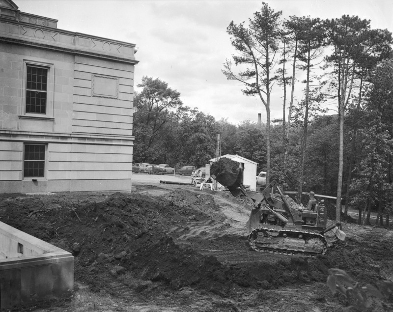 This 1957 photograph is of the beginning excavations for what will be the bookstore and Sun Room in the Memorial Union. A workman in a skid shovel with the shovel full of dirt and raised high waits and looks towards a dump truck that is emptying its load just beyond some trees. The earth around the skid shovel is stripped bare, tangled with tree roots and shows tire tracks under the raised shovel. The shovel has removed earth from near the terrace wall on the left. Two other workmen look at something on a table outside a white construction cabin beyond the back (south) corner of the Union.