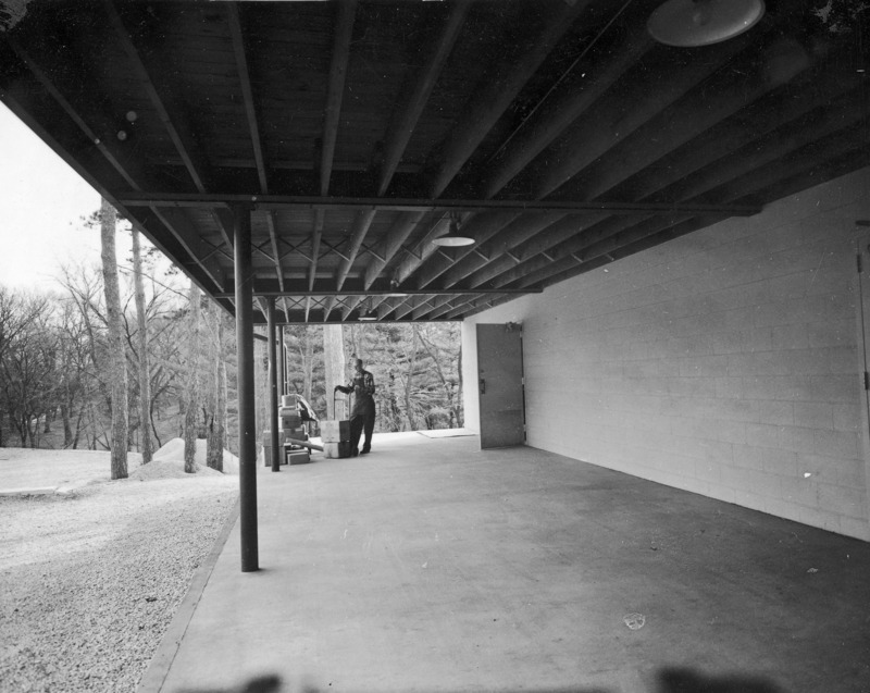 This 1958 photograph is of the loading dock behind the bookstore in the Memorial Union. A man with a hand truck is loading two boxes from a stack of freight at the far end of the dock. An open door to the Union building is behind him. Exposed roof joists and the support poles and beams frame the man and create perspective. Abundant trees fill the background.