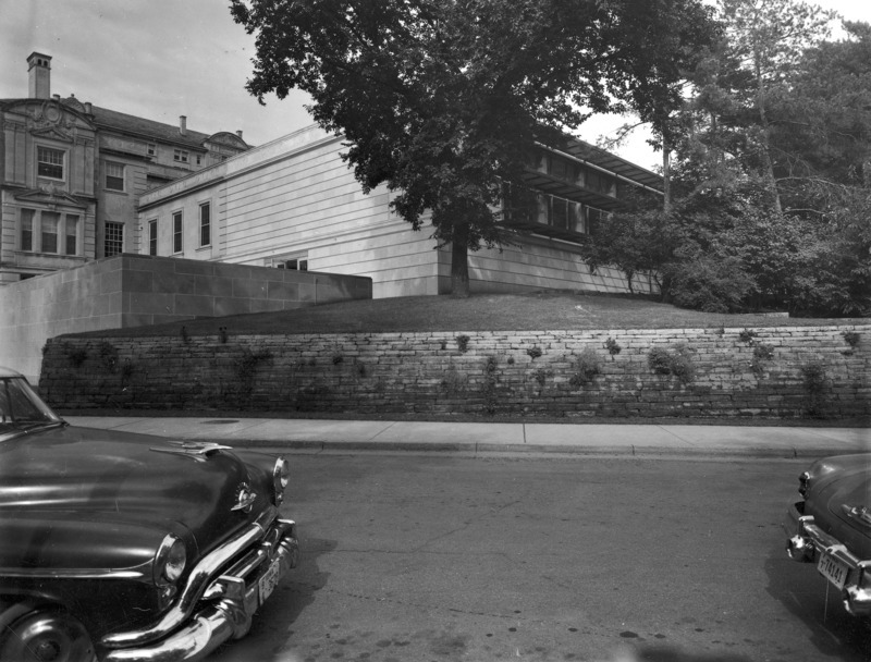 This 1958 photograph is of the Sun Room addition to the Memorial Union from street level. The front of one car and the back of another parked on the street are in the foreground. A stone retaining wall, the terrace wall, the walls of the Sun Room and the back (south) wall of the original building rise from the street. A large tree in the center of the photograph blocks most of the corner of the Sun Room.