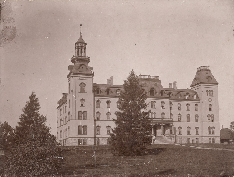 The front side of Iowa State's Old Main, with a large juniper tree blocking part of the view of the building. The Hub building can be seen to the extreme right of the image.