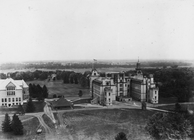 An aerial view of the rear side of Old Main. A flag flies from the north tower of the building. The Hub and railroad tracks are depicted to the left of Old Main. Further to the left of the image can be seen the rear of Morrill Hall.