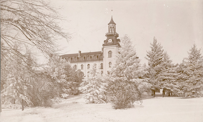 The south side of Old Main after a winter snow. A large number of trees and shrubs stand between the photographer and Old Main, partially blocking the view of the building.