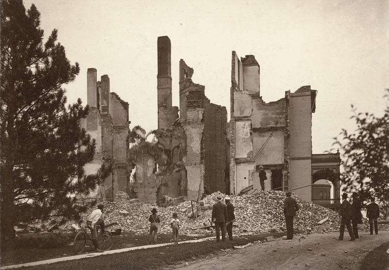 Old Main from the south after the second fire in August 1902. The gutted building stands behind a large heap of rubble, on which a man is standing. Seven men and two boys are in the foreground, all looking at the building remains.