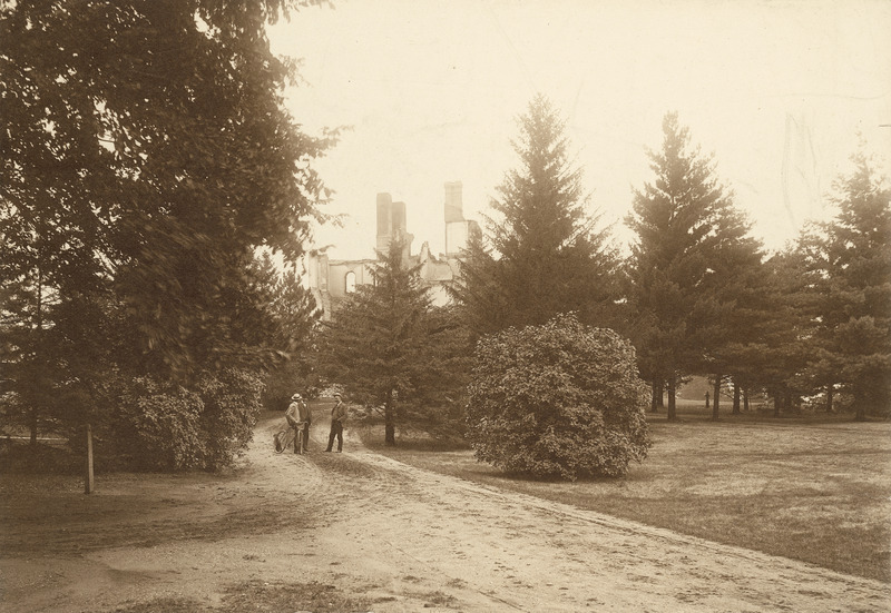 The burned remains of Old Main from the southeast after the second fire in August 1902. The gutted building stands behind a large grove of trees. Three men, one with a bicycle, stand talking in the middle of a drive that leads to the building.