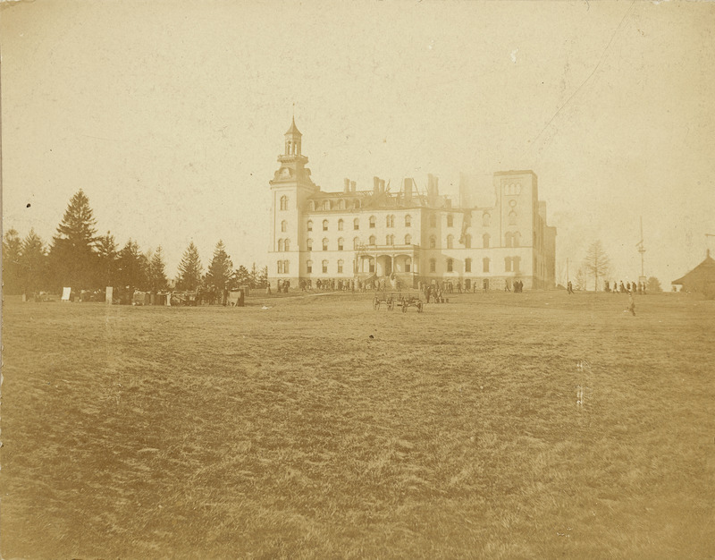 Old Main from the front showing the damage to the north side of the building after the first fire of December 1900. The Hub building located north of Old Main is also in the view. People are gathered in three different groups close to the building. A wagon sits on the lawn in front of the building.