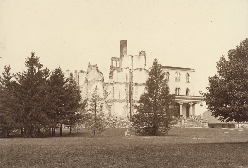 The remains of Old Main following the second fire in August 1902. An intact front porch looks out over a well groomed campus lawn. The Hub can be seen through a tree on the right of the image.