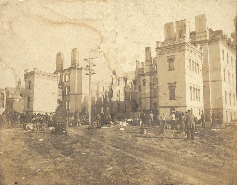 A rear view of the remains of Old Main following the first fire in December 1900. Room contents are scattered about the ground close to the building. A photographer is on the right of the scene with his back to the camera.