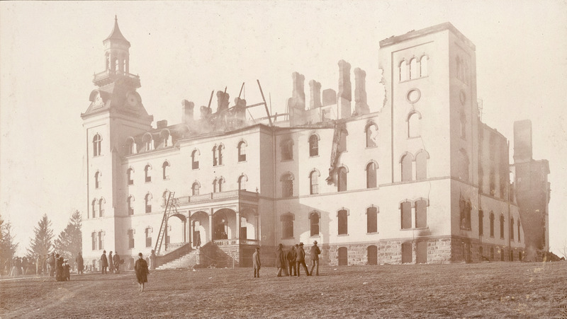 Old Main just after the first fire in December 1900. There is a little smoldering from the building center. The North wing has been greatly damaged. The roof appears to be gone entirely. People in coats are looking at the damage from the front lawn.