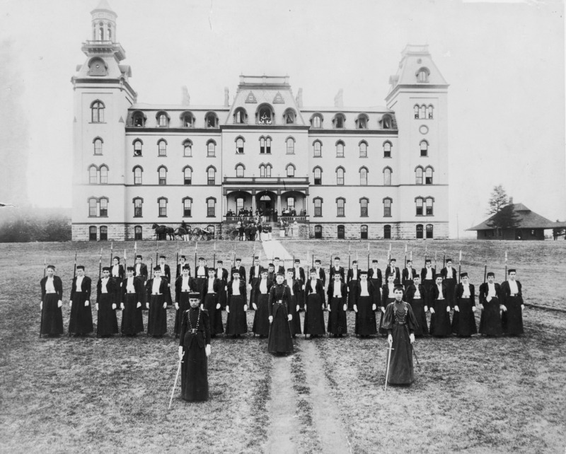 Old Main and the Hub building. A women's military corps (training occurred between 1878 and 1897) stands at attention in the foreground for the photograph. In the background, people are standing in front of the steps to Old Main and on the front porch. A wheeled cannon and a horse and carriage also are close to the entrance to Old Main.