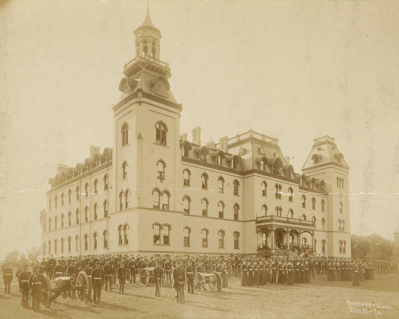 Old Main as the setting for a military corps review. Men's and women's corps are at attention. Military service for male students was mandatory between 1868 through 1962. For women, training was offered from 1878 through 1897. Three cannon are in the formation. An audience is on the stairs and front porch of Old Main.