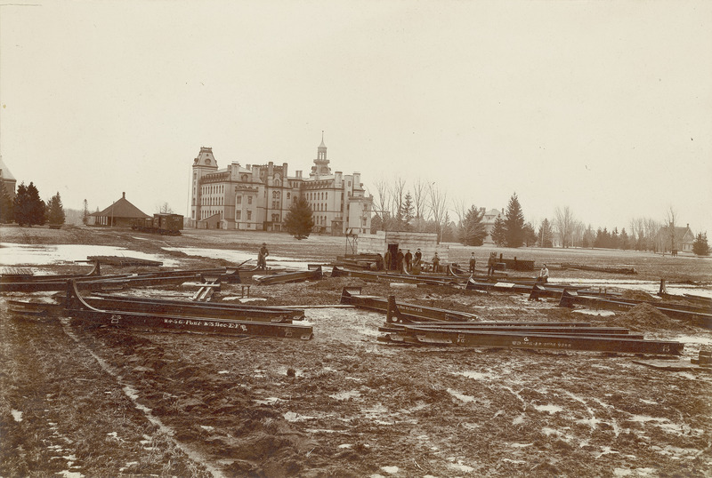 The Marston Water Tower in the early phases of construction. Old Main and a variety of other buildings appear in the background. Workmen stand around the site and there appears to be snow on the ground.