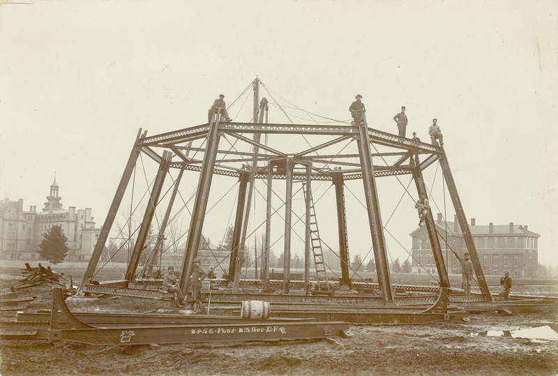 A view of the Water Tower construction site on March 18, 1897. Five of the construction crew members are sitting or standing on top of the first tier of support girders. Old Main and Chemistry Hall are among the buildings in the background.