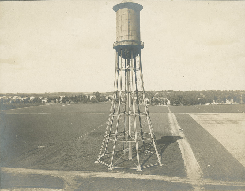 The Marston Water Tower from an elevated view. The Water Tower is surrounded by cultivated fields and dirt roads. Two people are walking on one of the roads, Other buildings and trees are in the distance.