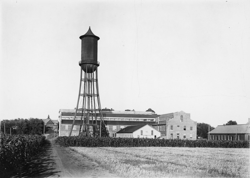 A field of corn and another crop surround the Marston Water Tower. Immediately behind the Water Tower is the Engineering Annex. A dirt road runs along the fields. In the distance is Morrill Hall and other buildings.