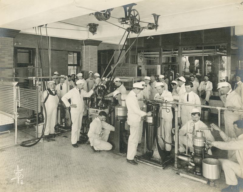 This January 10, 1905 photograph of dairy students shows students in a laboratory setting using milk separators and pasteurization equipment. The students are dressed in the traditional white jacket and trousers used in the dairy industry.