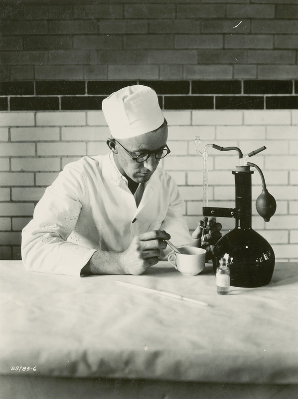 This photograph, taken July 16, 1930, shows E.F. Goss, Associate Professor of Dairy Industries at Iowa State College (University) using the Nafis Acid Test to measure the acidity of the milk, an important part of the process in making cottage cheese. Controlling the acidity of the milk as it sours keeps the curd firm, not mushy, and prevents the flavor of the cheese from becoming too sharp. Professor Goss is shown titrating a measured amount of milk in a white porcelain cup with an alkali solution that has been placed in a burette.