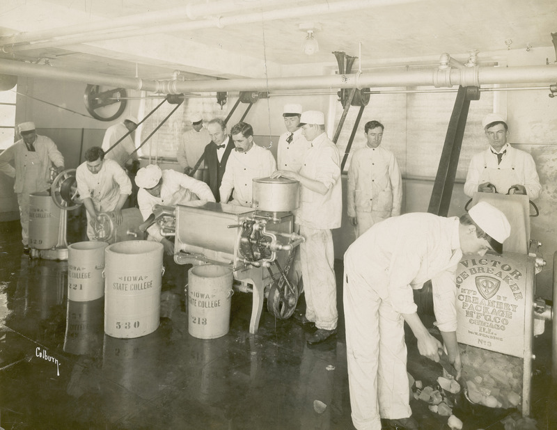 This photograph, taken about 1912, shows a group of students in the dairy laboratory using a power brine freezer for making ice cream. They are shown feeding cream into the machine at its right end, and waiting for it to be discharged into a large crock at the machine s left end. Two students at the right in the photograph are shown handling the ice needed in the ice cream making process. All of the students are dressed in the traditional white jacket and trouser uniform used in the dairy industry.