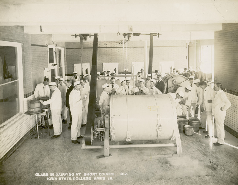 This photograph, taken in 1912, depicts a group of students in the creamery processing butter. They are shown working with motorized churns and wooden butter tubs. The churn agitates cream, bringing together its fat globules into lumps of butter, and leaving behind a liquid called buttermilk. The students are dressed in the traditional white jacket and trouser uniform used in the dairy industry.