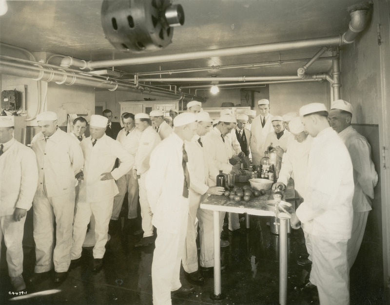 This photograph, taken in 1913 in the creamery, shows a group of students enrolled in the Iowa State College ice cream short course. Some students are working at a table with bottles of chemical reagents and pans. One of the students is whisking ingredients together in a pan. All of the students are dressed in the traditional white jacket and trouser uniform used in the dairy industry.