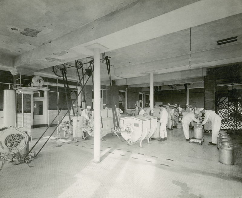 This photograph, taken in 1915 in the creamery, depicts a group of students processing butter. They are shown working with the motorized butter churns and wooden butter tubs. The churn agitates cream, bringing together its fat globules into lumps of butter, and leaving behind a liquid called buttermilk. A series of milk cans, used to transport milk from the milking parlor to the processing area, appear on the right. All of the students are dressed in the traditional white jacket and trouser uniform used in the dairy industry.