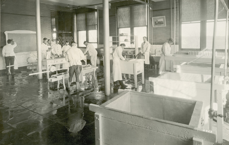 This photograph, taken in 1919, shows a group of students working in the cheese laboratory. Several cheese vats appear in the foreground. To the left in the photograph a student is standing by a cheese press, while several others are working at a table. All of the students are dressed in the traditional white jacket and trouser uniform used in the dairy industry.