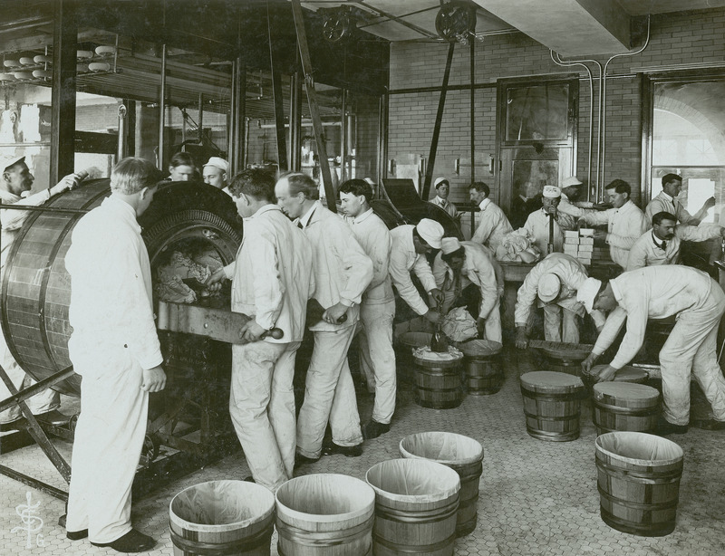 This photograph, taken in 1906 or 1907 in the Agricultural Annex, illustrates a group of students processing butter. They are shown removing butter from motorized butter churns and packing it into wooden tubs with liners. One student is leveling the butter in a tub while another seals a tub with a cover. All of the students are dressed in the traditional white jacket and trouser uniform used in the dairy industry.