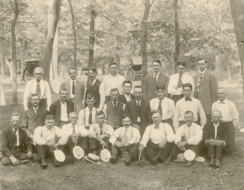 Members of the Benson Cow Testing Association are shown in 1909 in this photograph. The group is composed of twenty-four men. In the back row, third from the right is R.K. Bliss, who received a B.A. in animal husbandry from Iowa State College (University) in 1905, and was Director of Extension at Iowa State College (University), a position he held from 1914 to 1946. Several horse carriages of the phaeton/surrey style appear in the background.